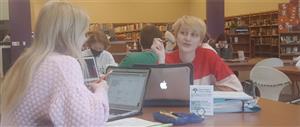 Students grouped up at tables in the library working on MacBooks and talking